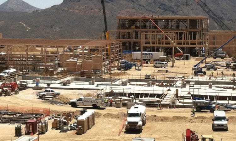 Construction site with several trucks, cranes, and workers; steel and wood frames are being erected and mountains are visible in the background.