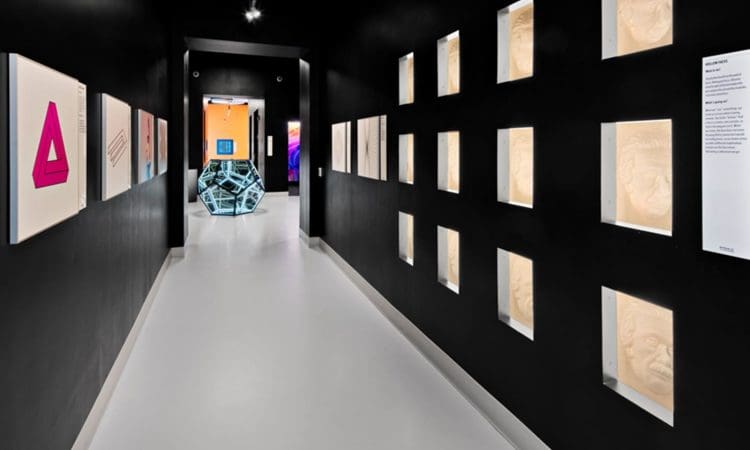 A modern hallway with a white floor, black walls displaying 3D face sculptures on the right, framed art on the left, and a geometric sculpture in a lit room at the end.