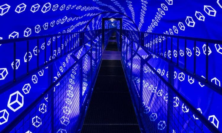 A walkway inside a tunnel with blue walls, featuring illuminated geometric cube patterns that create an optical illusion effect.