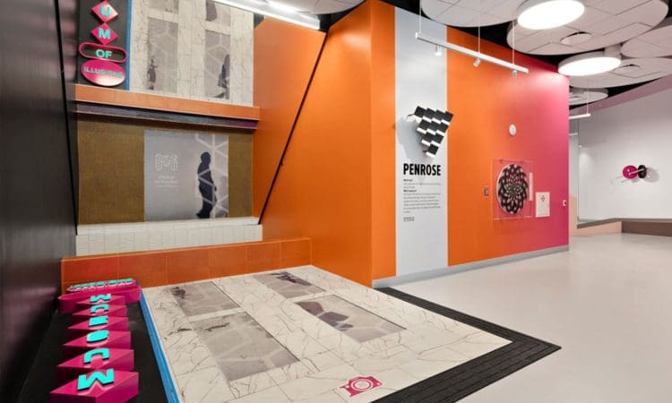 Modern museum exhibit with orange and white walls, geometric decor, informational panels, and interactive floor tiles spelling "MUSEUM." Ceiling features circular white panels.