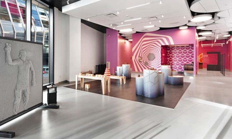 A modern exhibition space with geometric displays, a purple accent wall featuring optical art, and a large interactive wall sculpture of a human figure.
