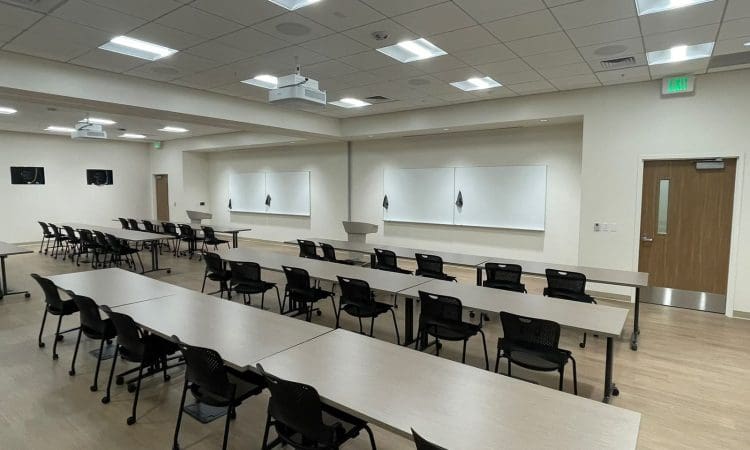 A modern classroom with rows of tables and chairs facing whiteboards and a podium, under bright ceiling lights. The room is empty.