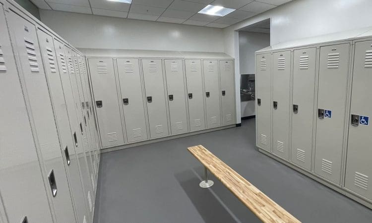A clean locker room with rows of beige metal lockers, a wooden bench in the center, and bright overhead lighting.
