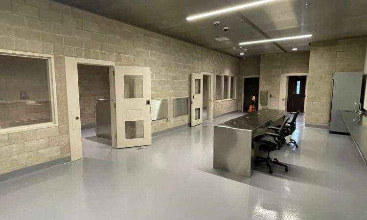 A clean, modern room with cinder block walls, several doors with windows, a metal table with office chairs, and large interior windows.