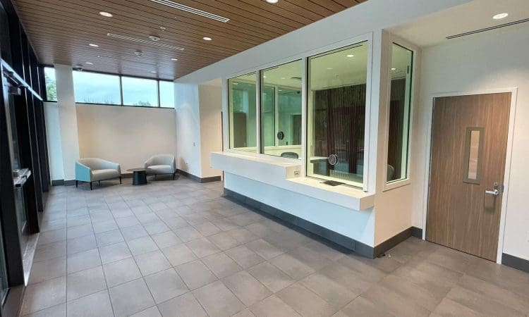 Modern waiting area with two chairs, a small table, tiled floor, wood-paneled ceiling, large windows, and a counter with glass partition next to a closed wooden door.
