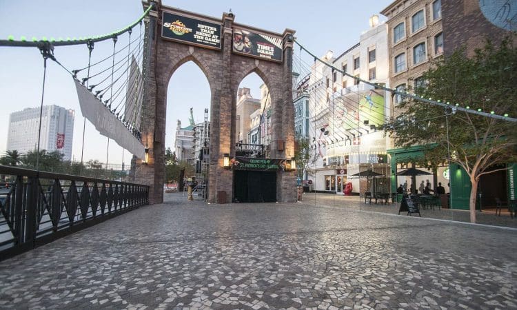 A stone arch bridge with overhead signs stands at the entrance to a pedestrian plaza lined with shops and buildings, under a clear sky.