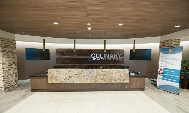 Reception desk at Culinary Health Center with four monitors, a marble front, wood accents, and a display banner on the right side.