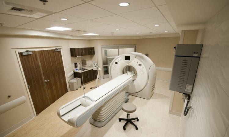 A hospital room with an MRI machine, control panels, a stool, cabinets, and double doors; the setting is clean and well-lit.