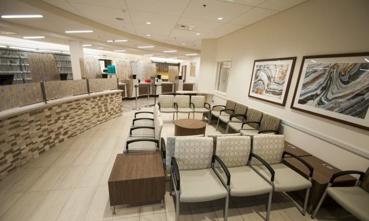 A modern, empty waiting room with rows of beige chairs, wooden tables, abstract artwork on the walls, and overhead lighting.