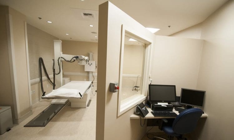 A medical imaging room with an X-ray machine on one side and a control station with monitors and a keyboard on the other side, separated by a window.