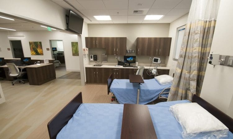 A hospital room with two beds, blue covers, bedside tables, medical equipment, cabinets, and a curtain divider; an adjacent nurse station is visible.