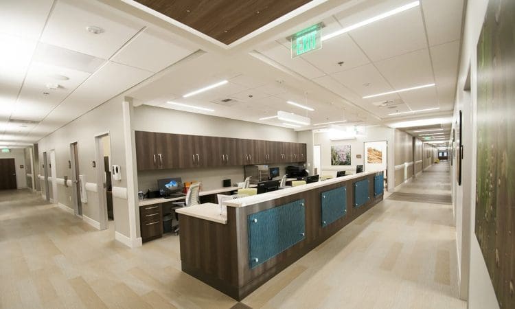 A modern hospital hallway with a central nurses' station, wood accents, computers, and bright overhead lighting. The corridor extends into the distance.