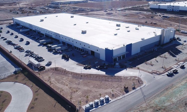 A large, white industrial warehouse with adjacent parking and loading docks, surrounded by undeveloped land and other industrial buildings.