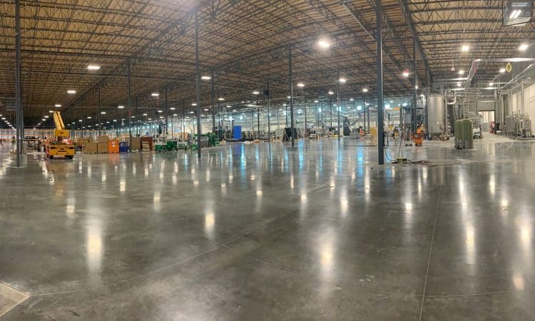 Wide view of a large, mostly empty warehouse with polished concrete floors, metal support beams, equipment, and storage containers scattered throughout. Bright overhead lighting illuminates the space.