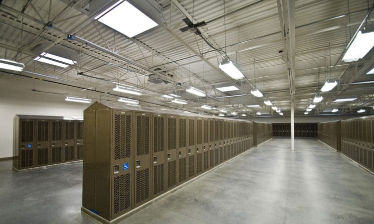 Rows of brown metal lockers line the walls of a large, brightly lit locker room with polished concrete floors and exposed ceiling fixtures.