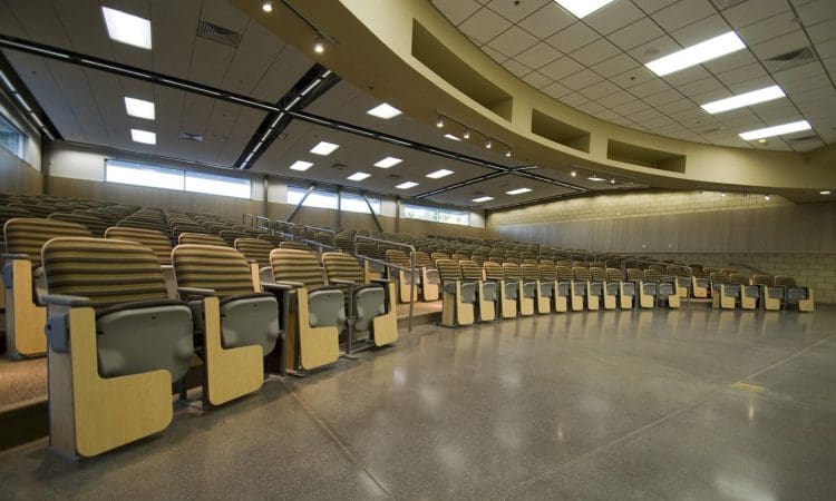 Empty lecture hall with rows of wooden seats and foldable desks, under a high ceiling with fluorescent lighting.