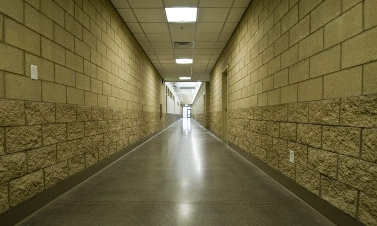 A long, empty hallway with tan concrete block walls, a polished floor, and fluorescent lights on the ceiling.
