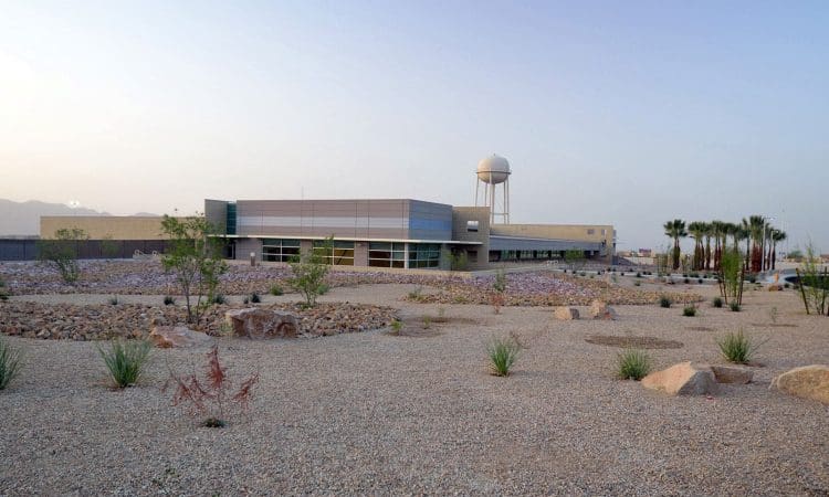 A modern low-rise building with large windows stands in a desert landscape with gravel, rocks, sparse plants, and a water tower in the background.