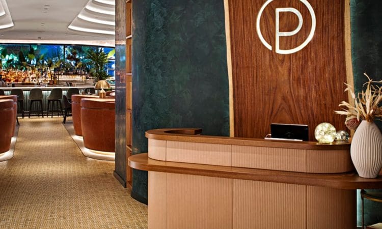 A modern restaurant entrance with a wooden reception desk, wall logo, and view of a bar and lounge area with leather chairs and decorative vases.