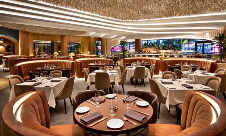 Spacious, modern restaurant with round and rectangular tables, tan upholstered chairs, set with plates and glasses, under a straw-textured ceiling and warm lighting.