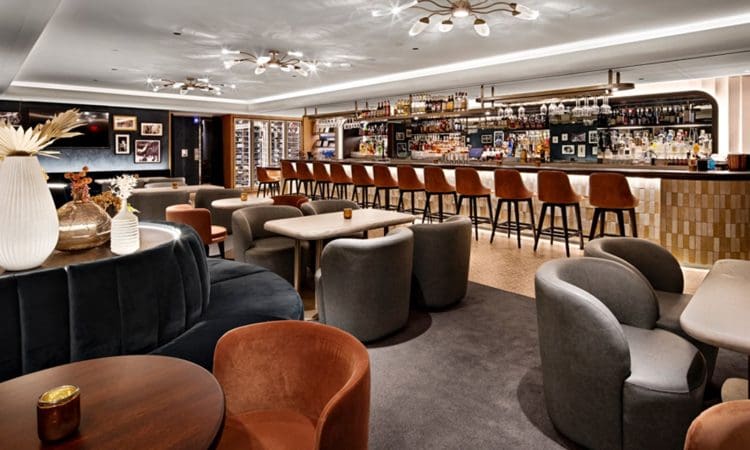 Modern lounge bar with plush seating, round tables, a long bar counter with stools, and decorative lighting fixtures on the ceiling.