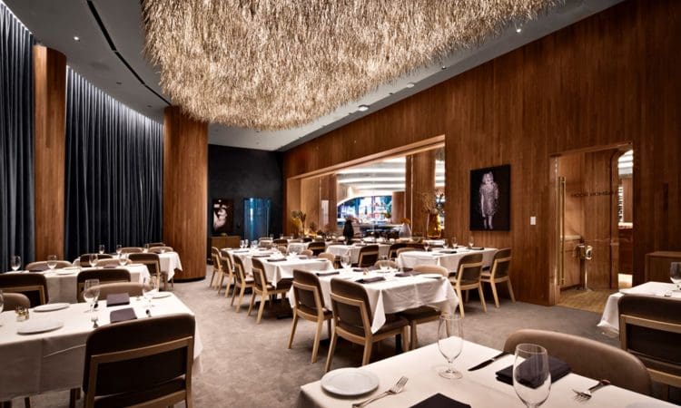 Modern restaurant dining room with wooden walls, white tablecloths, neatly set tables, upholstered chairs, and a large textured light fixture hanging from the ceiling.