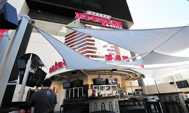 Outdoor bar and grill area with shaded canopy, barstools, flat-screen TVs, and a large "Rock Bar & Grill" sign above the entrance.