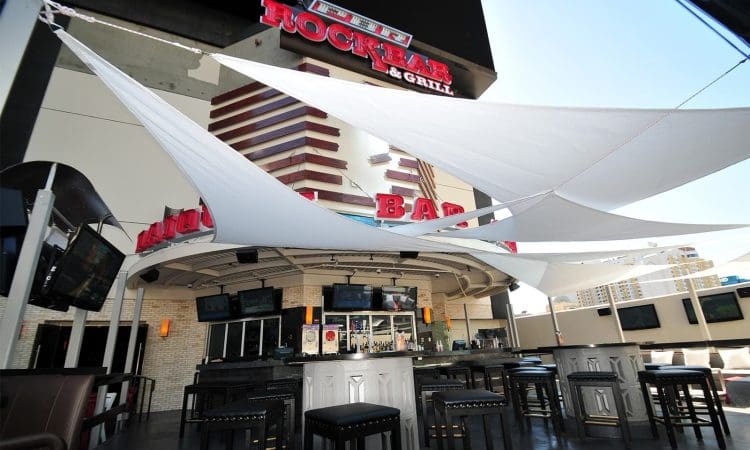 Outdoor bar area at PBR Rock Bar & Grill with high tables, barstools, several flat-screen TVs, and white shade sails overhead.