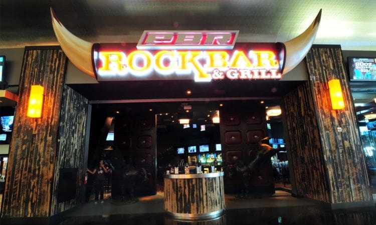 Entrance of a restaurant called "Rock Bar & Grill" with large illuminated signage, bull horns above the door, and wooden decor on the sides.