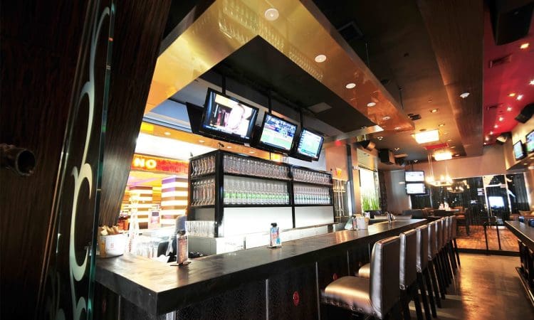 Modern bar interior with high chairs, multiple flat-screen TVs above the counter, glass shelves with bottles, and casino signage visible in the background.