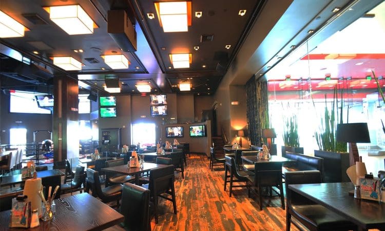 Modern restaurant interior with dark wooden tables and chairs, multiple TV screens on the walls, and large windows letting in natural light.