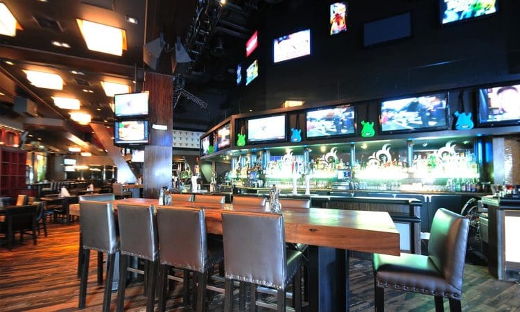 Modern sports bar interior with high tables and chairs, multiple TVs above the bar, illuminated shelves with bottles, and a wood-patterned floor.