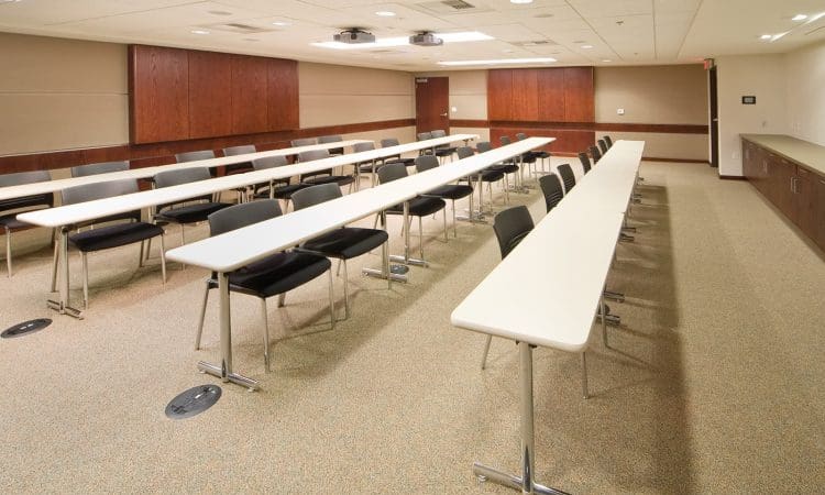 A modern classroom with long white tables, black chairs, beige carpet, wood paneling, ceiling lights, and a projector at the front; the room is empty.