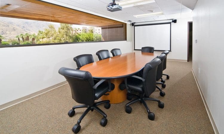 A conference room with a long wooden table, eight black office chairs, a projector, a projection screen, and a large window with a view of trees and mountains.