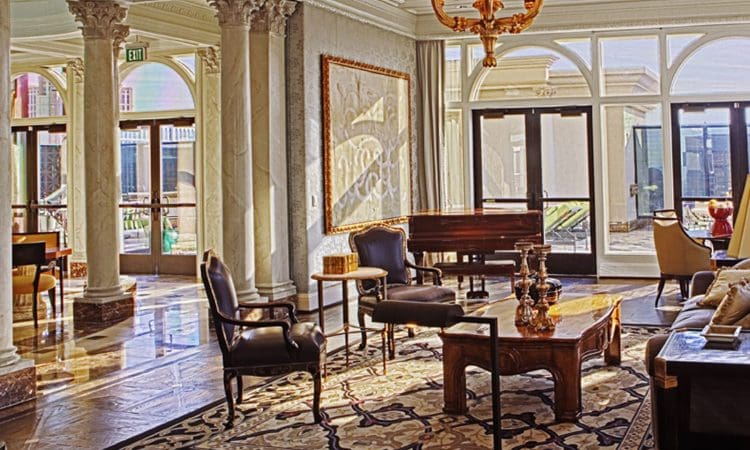 Elegant hotel lobby with marble columns, large windows, classic furniture, ornate rug, and a chandelier, lit by natural sunlight.
