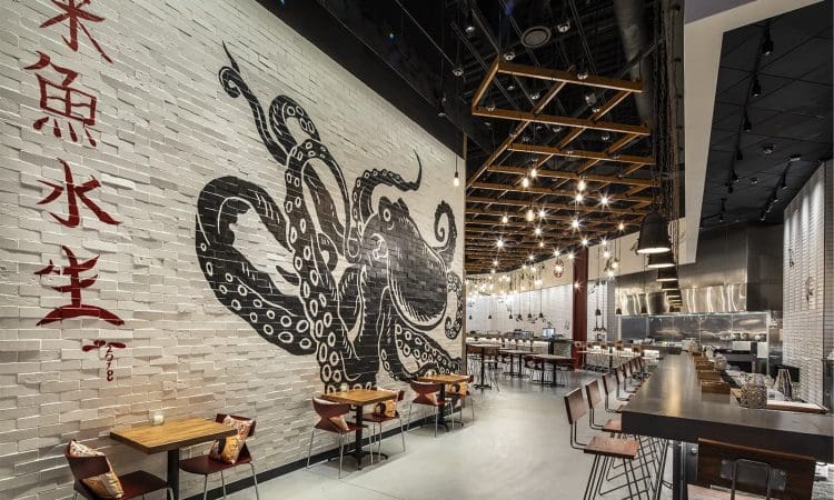 Modern restaurant interior with a large black octopus mural on a white brick wall, Asian characters, wooden tables, bar seating, and hanging lights.