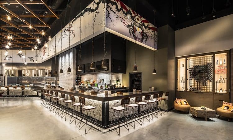 Modern restaurant interior with a central bar, high stools, open kitchen, wall art, and shelves displaying bottles; warm lighting and minimalist decor.