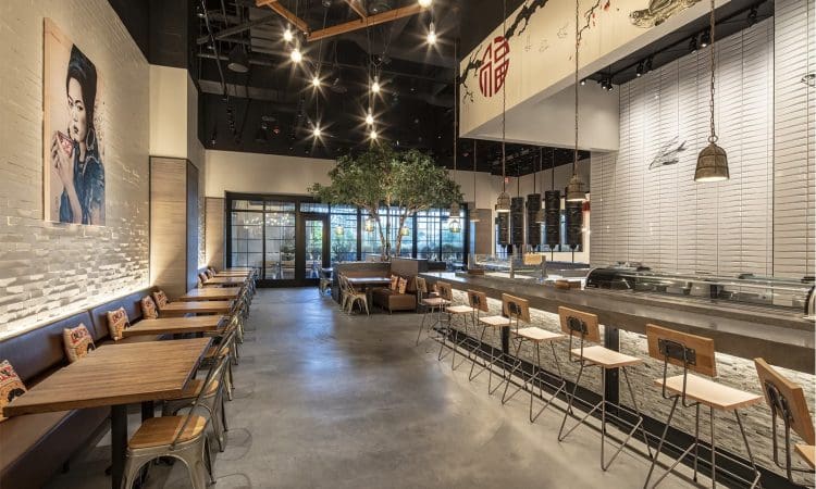 Modern restaurant interior with wooden tables, chairs, bar seating, wall art, and a central tree under ceiling lights. Large windows and minimalist decor create an open, airy atmosphere.