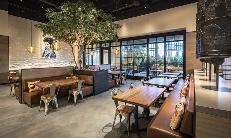 Modern restaurant interior with wooden tables, brown leather seating, metal chairs, a large indoor tree, pendant lights, and a wall mural of a woman in traditional attire.