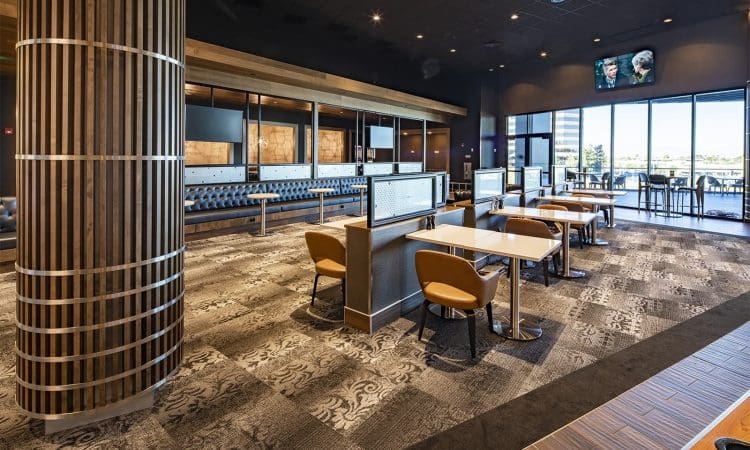 Modern restaurant interior with patterned carpet, wooden accents, booth and table seating, large windows, and a TV mounted on the wall.