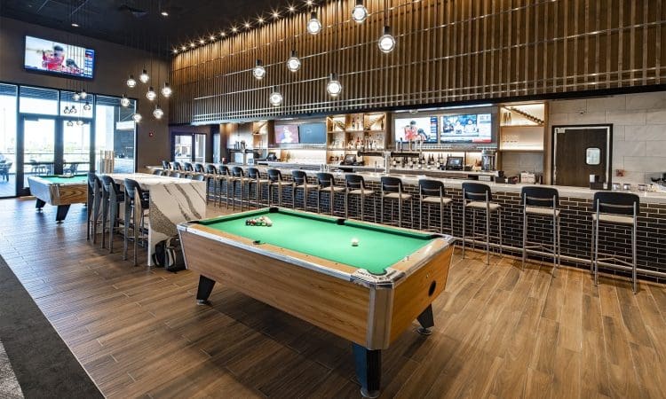 A modern bar with a long counter, barstools, multiple wall-mounted TVs, hanging lights, and a pool table in the center on a wood-patterned floor.