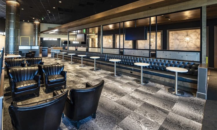 Modern lounge area with black leather chairs, a long tufted bench, round white tables, patterned carpet, and ambient lighting.