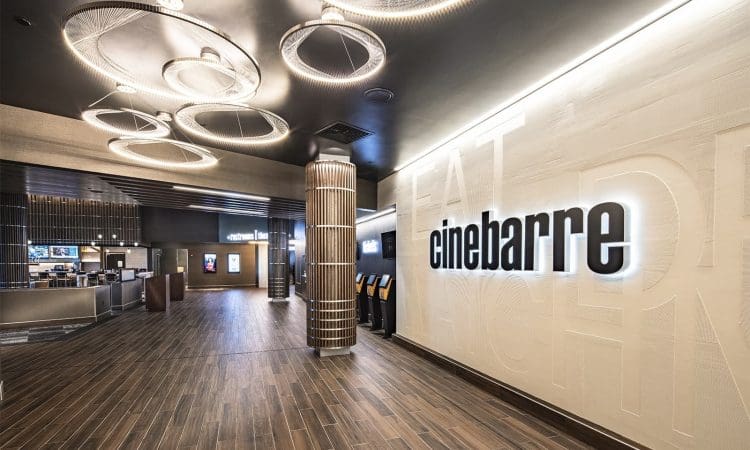 Modern movie theater lobby with wood-style flooring, illuminated ceiling lights, self-service kiosks, and a large "cinebarre" sign on a textured wall.