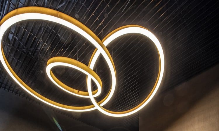Three large, circular, modern light fixtures with warm white illumination hang from a dark, slatted ceiling.