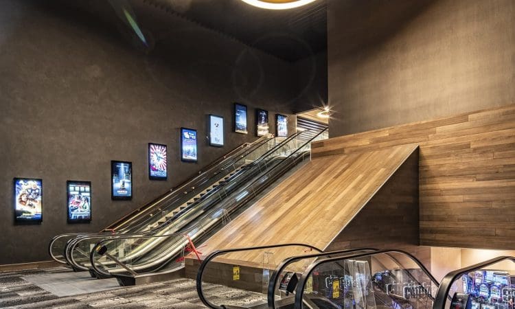 Modern indoor space with multiple escalators, digital movie posters on the wall, and a wood-paneled accent. The area is well-lit and appears to be part of a cinema or entertainment complex.