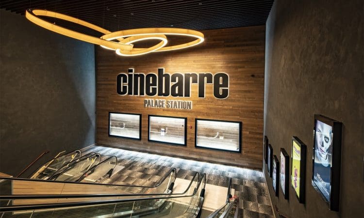 A modern escalator leads to the entrance of cinebarre Palace Station, with movie posters displayed on wood-paneled and gray walls.