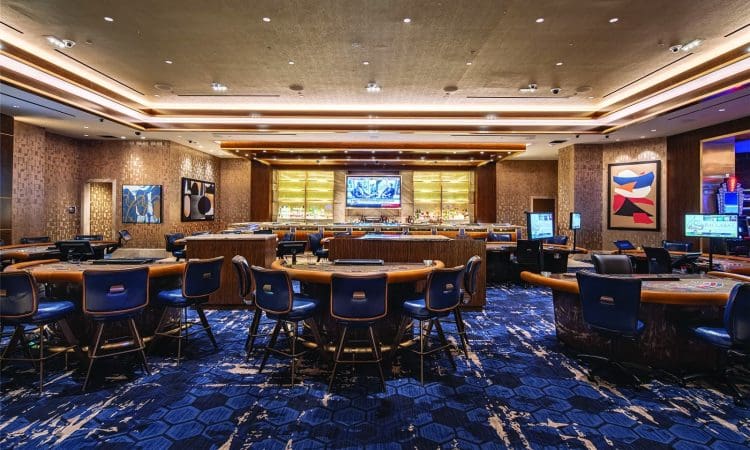 Spacious casino interior with gaming tables and chairs on a blue patterned carpet, modern lighting, wall art, and a large TV behind the bar area.