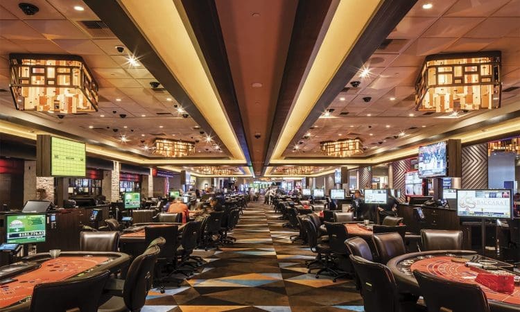 Spacious casino interior with rows of gaming tables and slot machines, illuminated by modern chandeliers, and people seated at various tables.