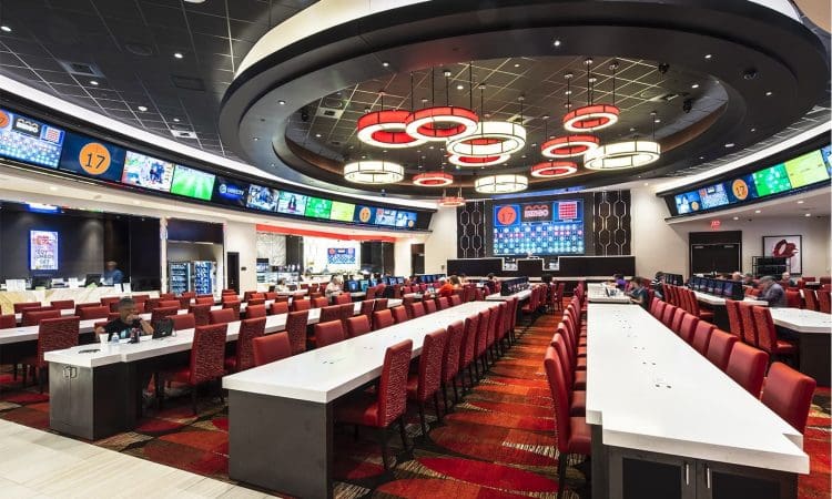 A spacious sports betting lounge with rows of red chairs, long white tables, large digital screens, and modern circular ceiling lights.