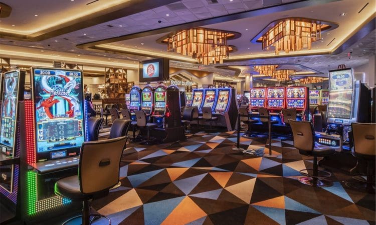 Casino interior with rows of colorful slot machines, patterned carpet, modern lighting fixtures, and empty chairs, creating a lively gaming atmosphere.